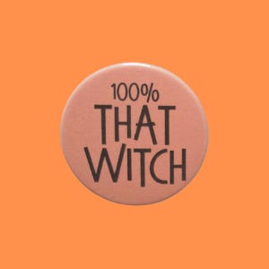 May include: A circular, pale pink button with the words "100% THAT WITCH" in black lettering. The button is set against an orange backdrop. A simple, bold statement.