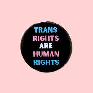 May include: A circular black badge with the words "TRANS RIGHTS ARE HUMAN RIGHTS" in blue and pink. The badge is set against a pale pink backdrop.