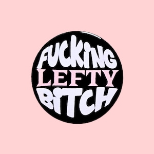 May include: A circular black badge with the text "FUCKING LEFTY BITCH" in white and pink. The badge is set against a pale pink backdrop.