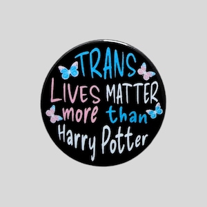 May include: A circular black button featuring the text "TRANS LIVES MATTER more than Harry Potter" in a mix of blue, pink, and white lettering. The design is accented with four small, colourful butterflies in shades of blue and pink.