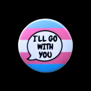 May include: A circular badge featuring the colours of the transgender pride flag: light blue, pink, and white. The badge has a speech bubble with the text "I'LL GO WITH YOU" in black.