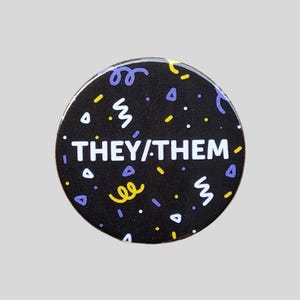 May include: Black button with a white background and the text "THEY/THEM" in white. The button is decorated with a confetti pattern in yellow and purple.