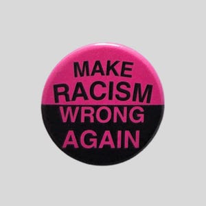 May include: A circular pin-back button featuring a horizontal split design. The top half is bright pink with the black text "MAKE RACISM" and the bottom half is black with the pink text "WRONG AGAIN" printed in a bold, sans-serif typeface.
