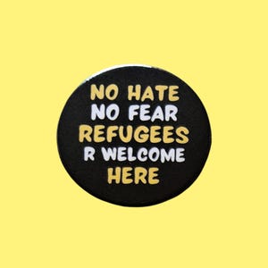 May include: A circular black badge with the words "NO HATE NO FEAR REFUGEES R WELCOME HERE" in white and gold. The badge is on a yellow background.