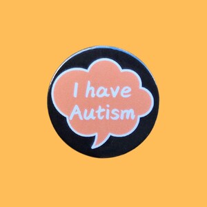 May include: A black and orange enamel pin with a speech bubble that says "I have Autism".