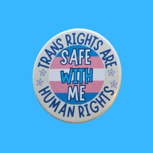 May include: A circular badge with the words "TRANS RIGHTS ARE SAFE WITH ME HUMAN RIGHTS" in blue and white. The design incorporates the transgender pride flag colours: pink, white, and blue, with star motifs.