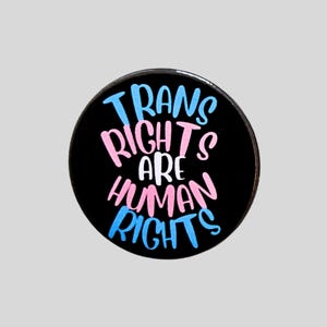 May include: A circular black badge featuring the words "TRANS RIGHTS ARE HUMAN RIGHTS" in blue and pink. The text is in a bold, clear font, set against a plain background.