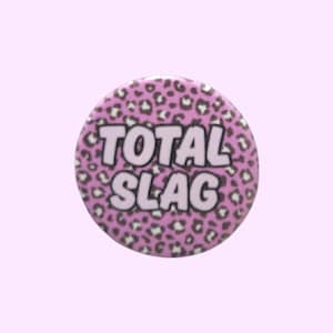 May include: A circular badge featuring a pink leopard print design. The words "TOTAL SLAG" are printed in white, bold letters across the centre. The background is a pale pink.
