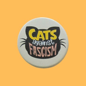 May include: A circular white pinback button featuring a black cat head silhouette. The text CATS is in yellow, against is in white, and FASCISM is in light pink. The design includes black whiskers extending from the sides of the cat head shape.