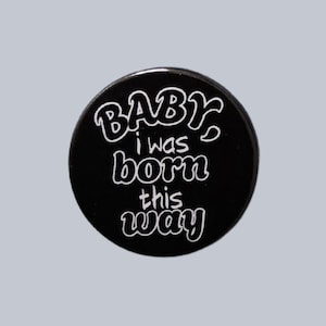 May include: A black button with the phrase "BABY I was born this way" in white, stylized lettering. The button is round and has a glossy finish. The text is centered on the button.