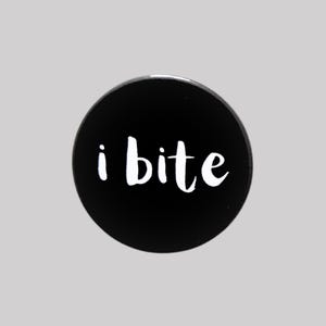 May include: A circular, black button featuring the white text "i bite" in a handwritten font. The button is set against a light grey backdrop.