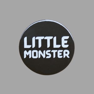 May include: A round, black button with the words "LITTLE MONSTER" in bold, white letters. The button has a white border and is set against a gray background. The design is simple and eye-catching.