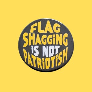 May include: A circular pinback button with a black background and yellow text that reads FLAG SHAGGING, with the words IS NOT in white, and the word PATRIOTISM in yellow at the bottom, featuring a bold, sans-serif, block-style font throughout.