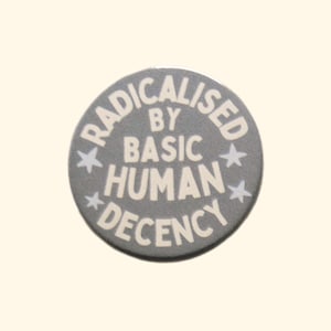 May include: A circular pinback button with a matte grey background and white text that reads RADICALISED BY BASIC HUMAN DECENCY. The text is arranged in a circular pattern around the centre, accented by four small white stars placed between the words.
