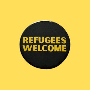 May include: A circular black badge with the text "REFUGEES WELCOME" in yellow capital letters. The badge is set against a yellow backdrop. The design is simple and direct in its message.