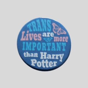 May include: A circular blue pinback button featuring a pink butterfly graphic and the text: Trans Lives are more IMPORTANT than Harry Potter. The text is displayed in a mix of light blue, pink, and white lettering against a dark blue background.