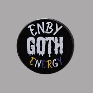 May include: A round, black button with the words "ENBY GOTH ENERGY" in white, yellow, and purple lettering. The word "GOTH" has a dripping effect. The button is set against a gray background.