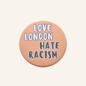 May include: A circular badge in a peach hue, featuring the text "LOVE LONDON HATE RACISM" in white and black. The design is straightforward and the finish is matte.