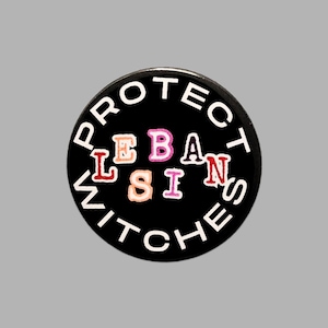 May include: A circular black pin-back button featuring the white text PROTECT WITCHES arranged in a circle around the centre. The word LEBIANS is printed in the middle using multi-coloured, textured letters in shades of pink, orange, and red.