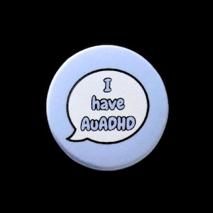 May include: A circular, pale blue badge featuring the text "I have AuADHD" within a white speech bubble, outlined in black. The badge is set against a black backdrop.
