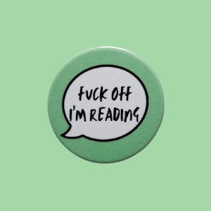 May include: A circular, pale green badge featuring a white speech bubble with the text "FUCK OFF I'M READING" in black, handwritten font. The badge has a black outline and is set against a light green backdrop.