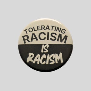 May include: A circular pinback button divided horizontally into two halves. The top half is off-white with the black text TOLERATING RACISM. The bottom half is black with the off-white text IS RACISM. The design features a bold, high-contrast look.