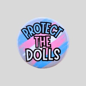 May include: A round button with the text "PROTECT THE DOLLS" in black outlined blue letters. The background has pink, blue, and white stripes. The button is a light gray color.