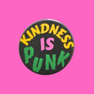 May include: A circular pin-back button with a black background. The text "KINDNESS" is printed in yellow, "IS" in pink, and "PUNK" in green. The letters are arranged in a curved fashion around the centre of the circular button surface.