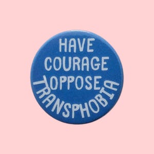 May include: A circular blue pinback button featuring white text that reads HAVE COURAGE OPPOSE TRANSPHOBIA. The text is arranged in four lines, with the final word curved along the bottom edge of the button against a solid blue background.
