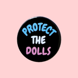 May include: A black button with the text "PROTECT THE DOLLS" in blue, white, and pink lettering.