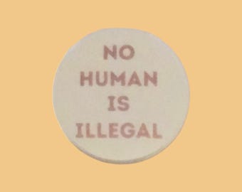 May include: A round, off-white object with the words "NO HUMAN IS ILLEGAL" printed in a reddish-brown color. The object is centered on a light orange background.