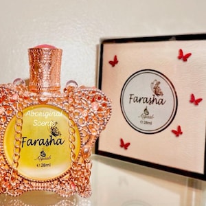 May include: A gold and pink perfume bottle with a crown-shaped design and a butterfly on the label. The bottle is labeled "Farasha Aboriginal Scents e 28ml". The bottle is sitting on a white surface with a gold and pink box behind it.