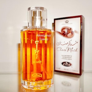 May include: A clear glass perfume bottle with a gold-colored liquid and a clear cap. The bottle has the text "Choco Musk" and "Aboriginal Scents" on it. A box with the text "Choco Musk" and a crown logo is behind the bottle. The bottle contains 50 ml.