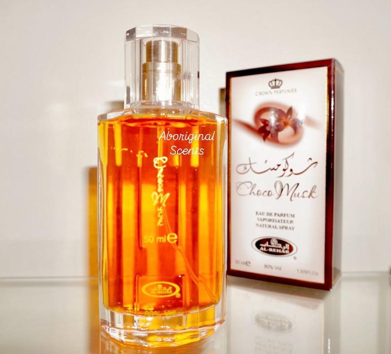 May include: A clear glass perfume bottle with a gold-colored liquid and a clear cap. The bottle has the text "Choco Musk" and "Aboriginal Scents" on it. A box with the text "Choco Musk" and a crown logo is behind the bottle. The bottle contains 50 ml.
