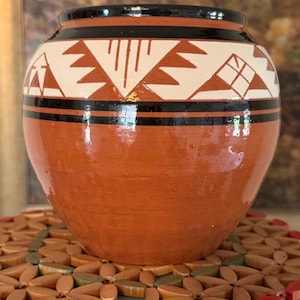 Op de afbeelding: Een terracotta-kleurige keramische pot met een ronde vorm en een brede opening. Het bovenste gedeelte is voorzien van een band met witte geometrische patronen en strakke zwarte lijnen op de natuurlijke kleibasis, wat een duidelijk contrast geeft.