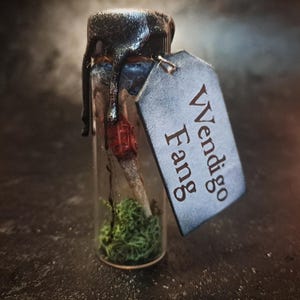 May include: A small glass vial with a black wax seal and a tag that reads "Wendigo Fang." Inside the vial is a bone-like object with a red tip, surrounded by green moss. The vial is sealed with black wax.