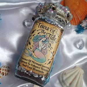 May include: A glass bottle with a silver lid and a label that reads "Tears Of The Tide". The label features a mermaid with pink and blue hair and a tail. The bottle is filled with a purple liquid and glitter.