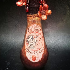 May include: A small, pear-shaped glass bottle with a gold metallic finish. The bottle has a label that reads "Eternal Drops of Love" and features a black and white illustration of a woman holding a heart. The bottle is decorated with red and orange flowers.