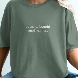 Peut inclure: Un t-shirt à manches courtes vert sauge avec le texte blanc "oops, I bought another hat" imprimé dans une police de style machine à écrire au centre de la poitrine. Le t-shirt présente une encolure ronde et une coupe ample et décontractée.