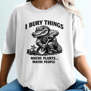 May include: White t-shirt with a humorous graphic. The design features an alligator character digging with a shovel, accompanied by the text "I BURY THINGS" above and "MAYBE PLANTS... MAYBE PEOPLE" below. The shirt is a casual, unisex style.