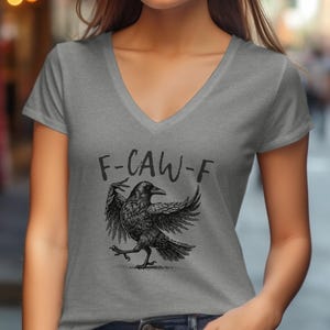 Puede incluir: Camiseta gris con cuello en V con una ilustración de un cuervo negro y el texto "F-CAW-F". El cuervo se muestra con las alas extendidas, dando a la camiseta un aspecto único y artístico. El diseño es detallado y llamativo.