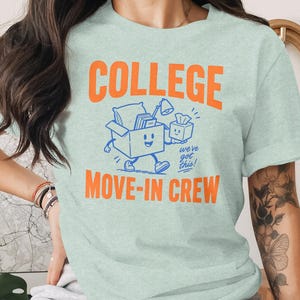 May include: A light green t-shirt featuring the orange text COLLEGE MOVE-IN CREW. Below the word COLLEGE is a blue graphic of a smiling cardboard box with arms and legs, carrying a pillow, books, a lamp, and a tissue box, with the text we've got this!