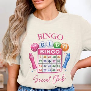 May include: Cream-colored t-shirt with a Bingo theme. The design includes the word "BINGO" in pink, along with bingo balls and a bingo card. The text "Social Club" is also present. Two pink and blue markers are on either side of the bingo card.