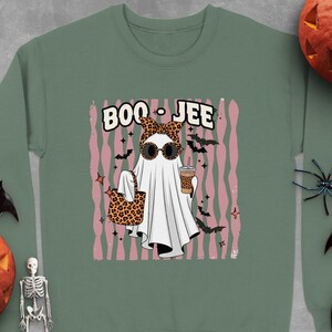 May include: A sage green crewneck sweatshirt featuring a graphic of a white ghost wearing a leopard print headband and sunglasses. The ghost holds a leopard print tote bag and a coffee cup. The text BOO-JEE is printed above the ghost.