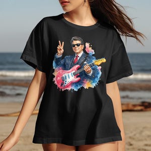 Person wearing black t-shirt with watercolor portrait of guitarist playing pink guitar.