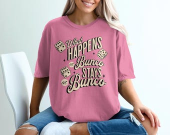 Könnte beinhalten: Ein pinkfarbenes T-Shirt mit dem Text "What Happens at Bunco Stays at Bunco" in einem Vintage-Schriftstil, begleitet von Würfelgrafiken. Das Shirt hat einen lässigen, übergroßen Stil.
