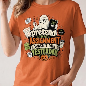 May include: An orange t-shirt featuring a graphic of a ghost wearing glasses, holding a red pen and a grade book. The text reads: pretend THAT ASSIGNMENT WASN'T DUE YESTERDAY. Additional graphics include a pumpkin, spiderwebs, and a coffee cup.