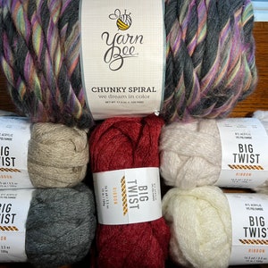Op de afbeelding: Zes bollen garen in verschillende kleuren, waaronder grijs, beige, wit en rood. Het garen is geëtiketteerd "Big Twist Ribbon" en is gemaakt van acryl en polyamide. Het garen is 14,5 yards per bol.