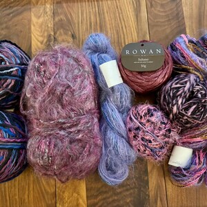 Op de afbeelding: Een verzameling kleurrijke garenbollen in verschillende texturen en tinten paars, blauw en roze. Sommige bollen zijn strak gewikkeld, andere zijn pluizig en getextureerd. Eén bol heeft een label met de tekst "ROWAN Sultano 50g".