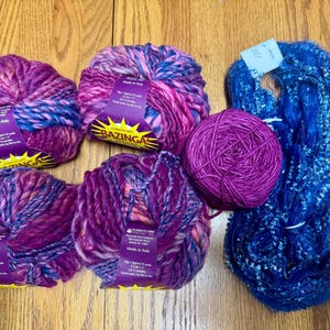 Op de afbeelding: Vier bollen garen in tinten van roze, paars en blauw, met een bol effen paars garen. Het garen is geëtiketteerd met "Bazinga!" en "Made in Italy".